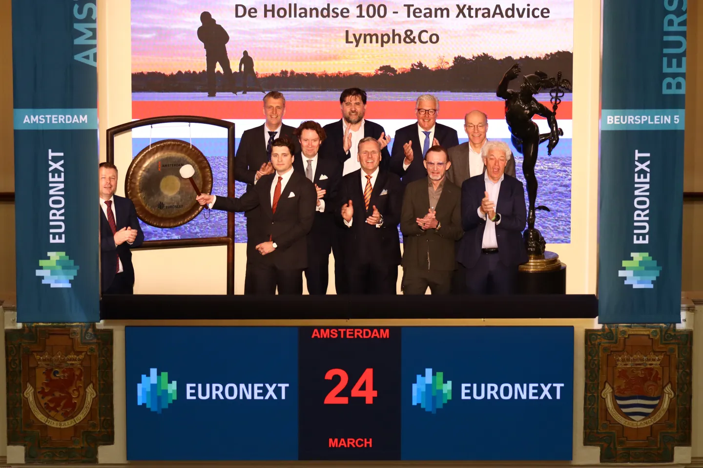Representatives from XtraAdvice and Lymph & Co stand together as the ceremonial gong is rung at Beursplein 5, marking the start of the 10th Hollandse 100 and honouring the memory of Rudolf van Schaik.